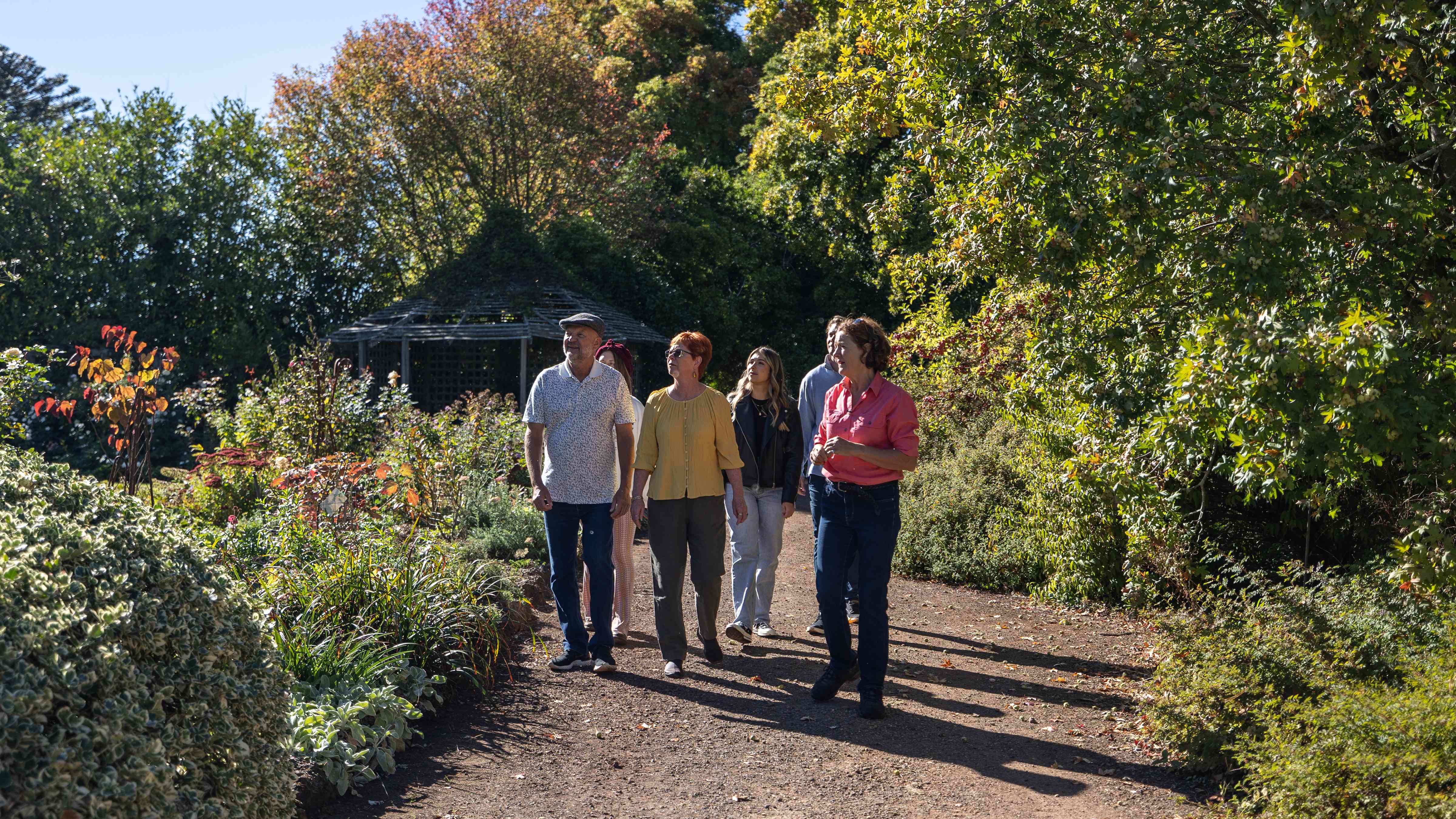 A family group is wandering along a driveway being guided by a member of the Archer family. The driveway is bordered with an array of shrubs on the left including a Forest Pansy, roses, penstemons, and a varigated, and a Malus trilobata tree on the right with a timber summer house and larger trees in the background. Photo: Kate von Stieglitz / Tourism Australia.