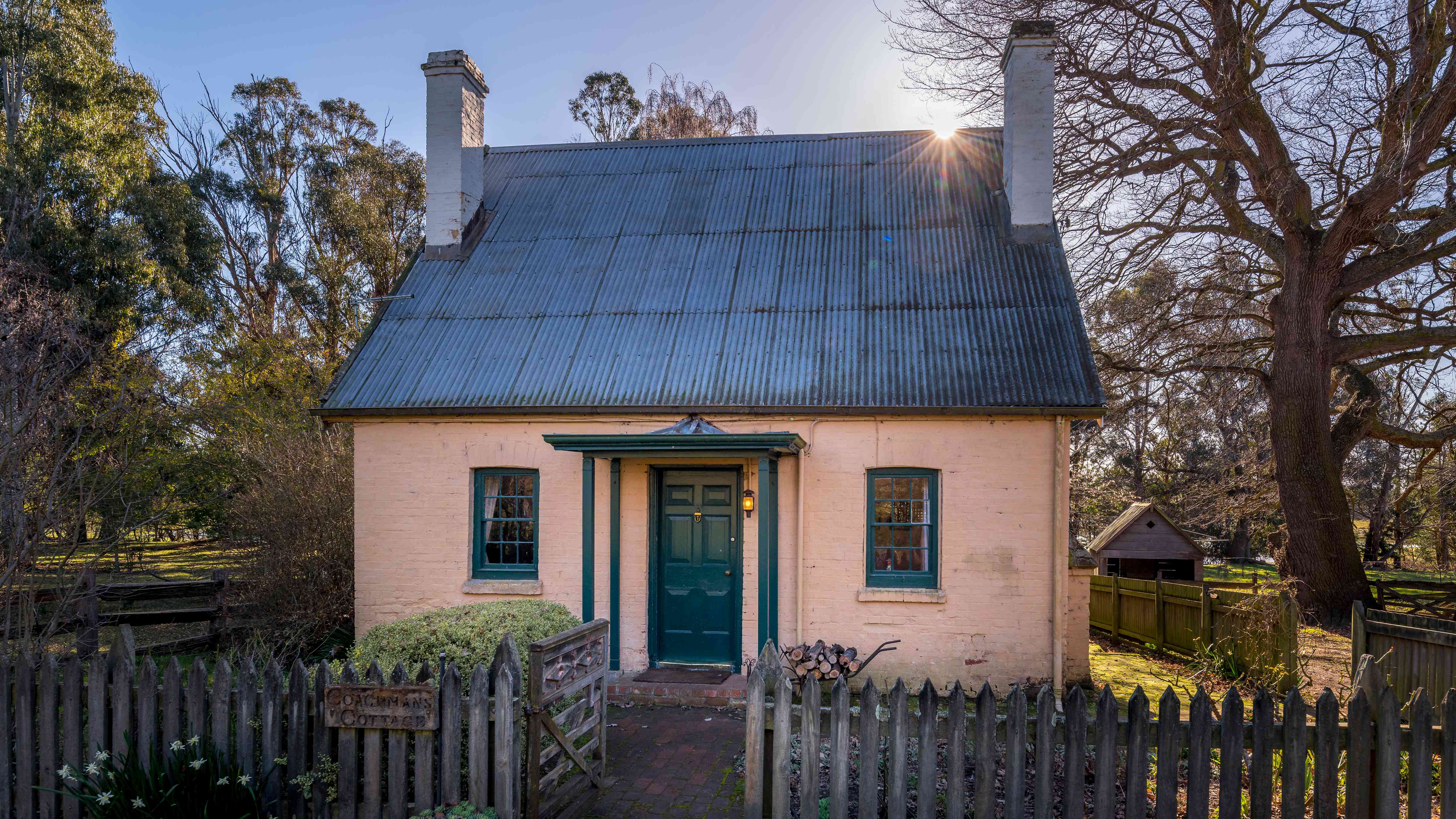 A Georgian cottage with a steep pitched corrugated iron roof, two chimneys and two georgian paned windows on either side of the front door which has a small porch over. The walls are brick and painted with a pale pink limewash. A picket fence is in the foreground with a gate open leading to the cottage. A large Oak tree stands to the right of the cottage and gum trees in the background. Photo: Rob Burnett.