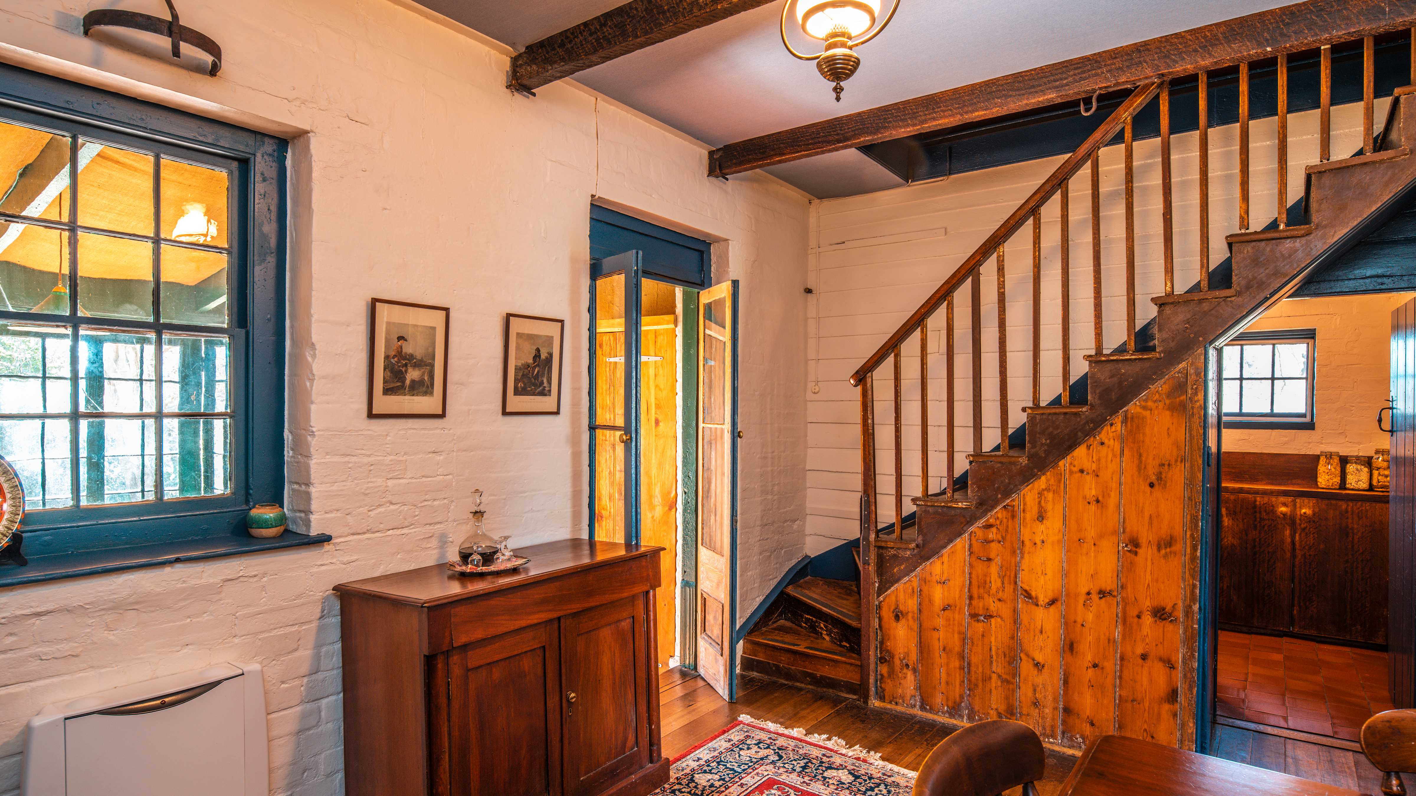 The dining room has an antique cupboard with a decanter of port and glasses sitting on it. A door and window are on the left of the image and a staircase with a timber railing goes up to the second floor. Another door is open into the kitchen. Timber boards on the floor are covered by a Persian rug. Photo: Rob Burnett.