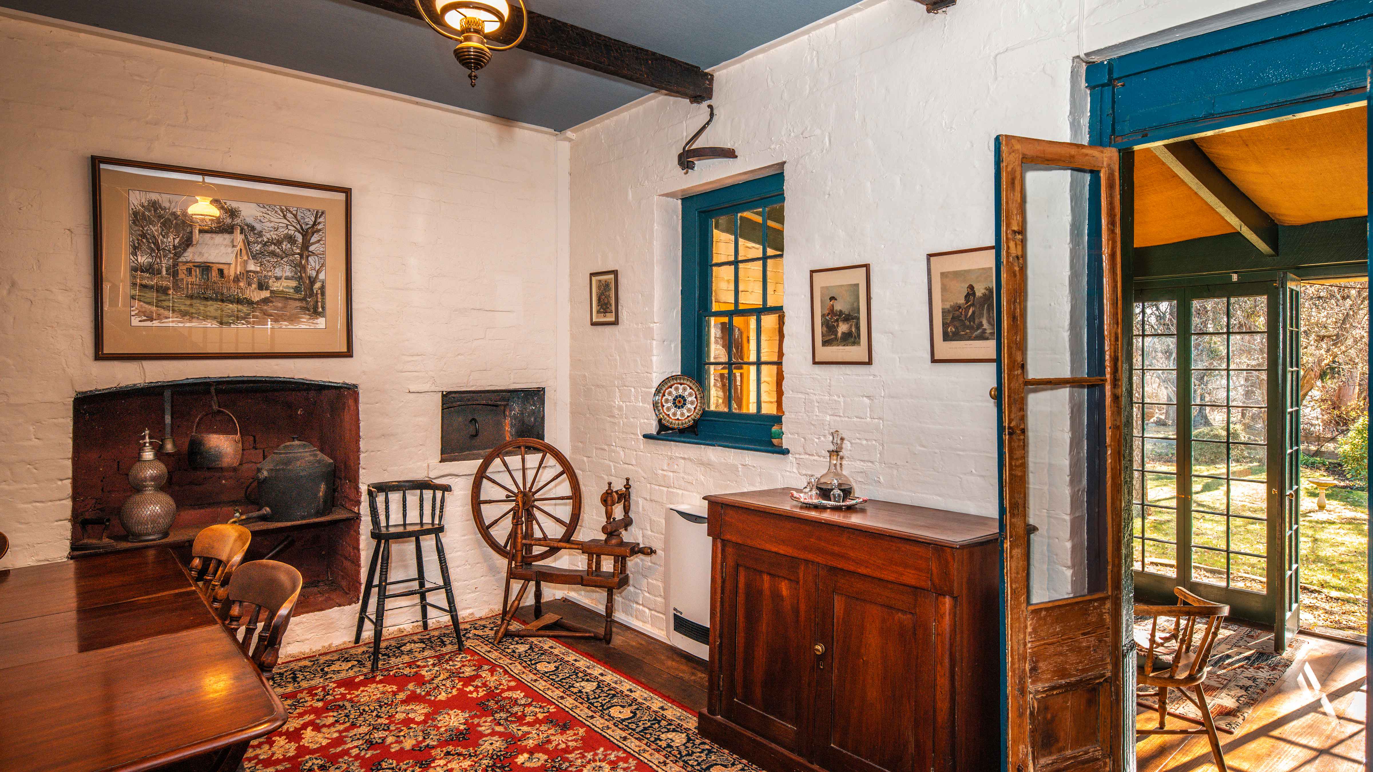 The dining room has a antique timber table and chairs on the left with numerous antiques on display including a glass soda syphon, cast iron water heater with tap, child’s highchair, spinning wheel and a cedar sideboard with a decanter of port and glasses on a silver tray. A Persian rug cover timber floor boards and a door with glass panes is open to the sunroom with a further door open to the garden in the background. Photo: Rob Burnett.