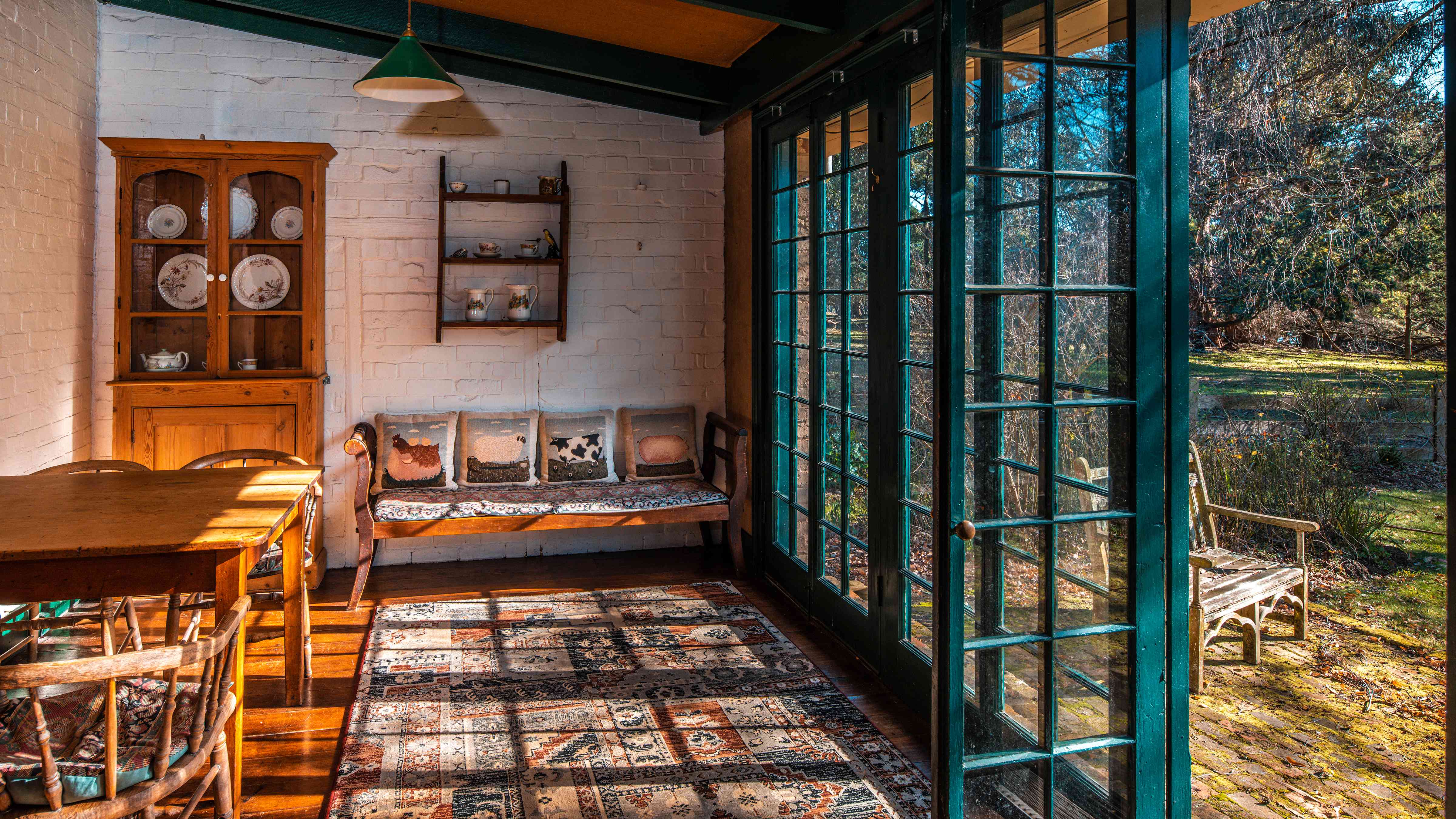 A timber table with antique chairs stands to the left of the image with a pine dresser behind. The dresser along with a set of wall shelves has china plates and jugs displayed. An antique timber settee has four farm animal print cushions standing up against the wall and a persian patterned rug is on the floor. A wall of glass paned doors look out to the garden with a timber chair standing on brick paving. Photo: Rob Burnett.