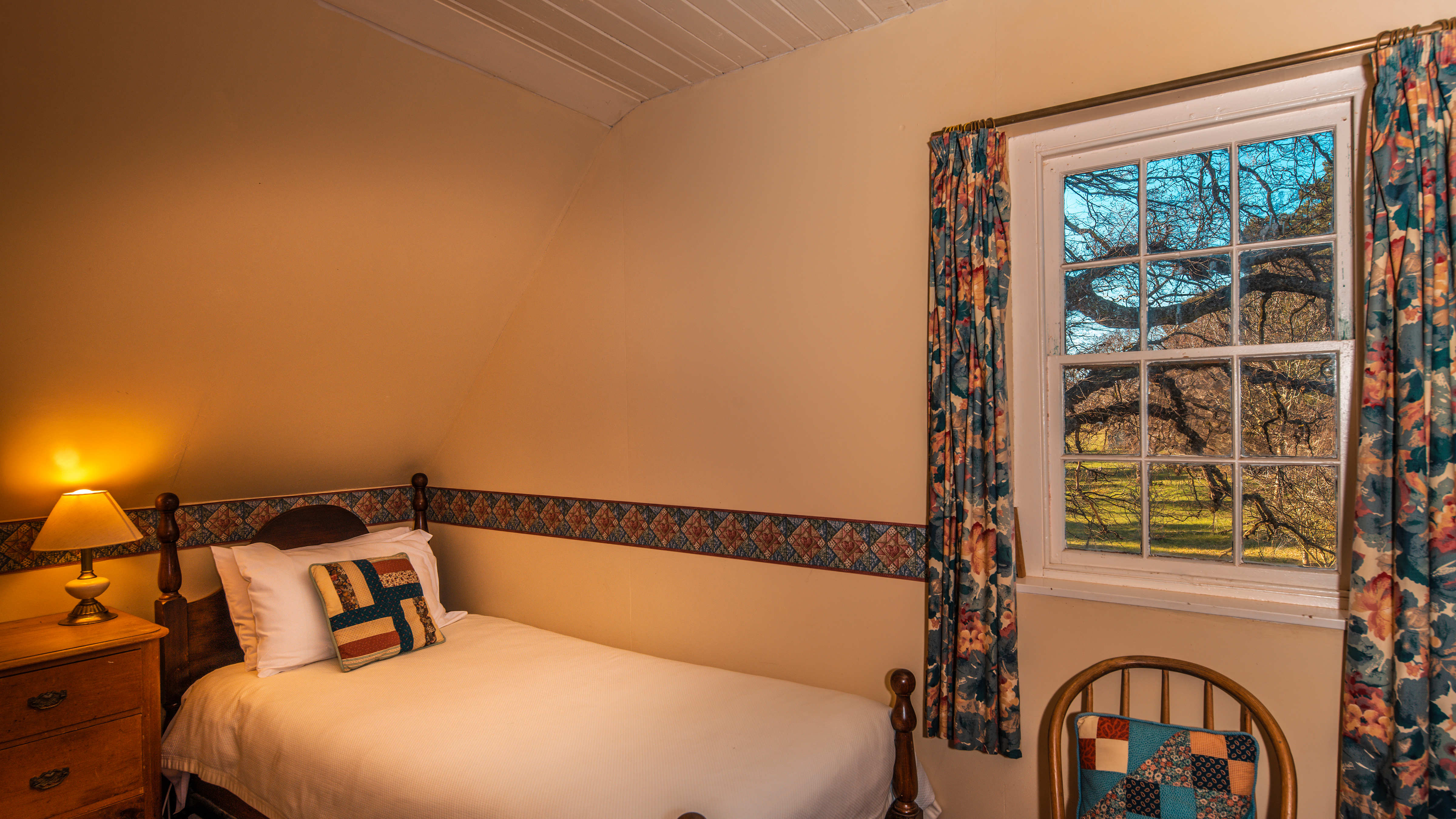 The room has a sloped ceiling and a paned window looking out to an Oak tree. The single timber bed is made with a white doona, white pillows and a cream,blue and maroon patterned cushion. There is a decorative freeze on the wall and a bedside lamp turned on and sits on a pine chest of drawers. A wooden chair sits under the window which has patterned curtains pulled to each side. Photo: Rob Burnett.