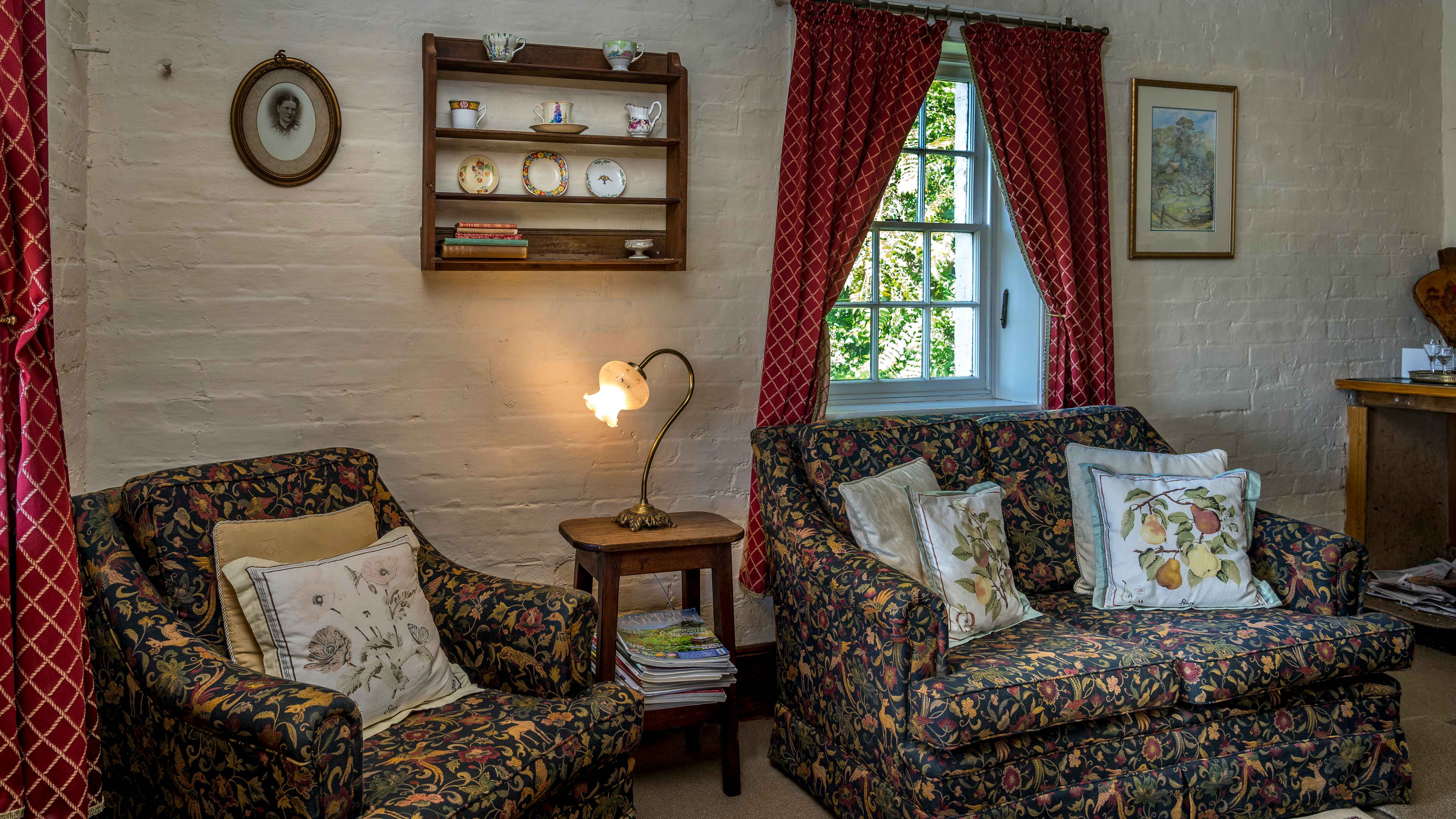 A single lounge chair and couch, covered with a patterned fabric, have pale patterned cushions on them. A wooden stool has an antique brass lamp on with magazines under. A small set of wooden shelves hangs on the wall with tea cups, saucers and books on the shelves. A couple of pictures hang on the wall and a window with red curtains looks out to green foliage. Photo: Rob Burnett.