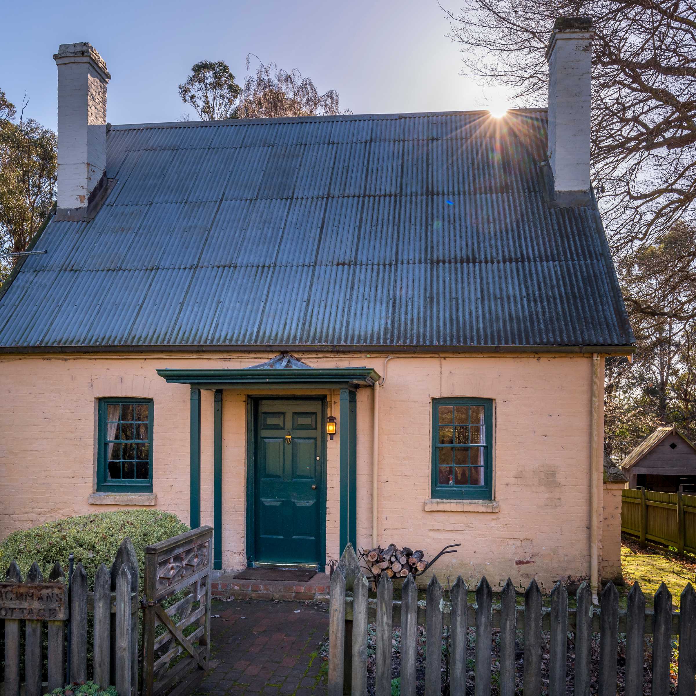 A Georgian cottage with a steep pitched corrugated iron roof, two chimneys and two georgian paned windows on either side of the front door which has a small porch over. The walls are brick and painted with a pale pink limewash. A picket fence is in the foreground with a gate open leading to the cottage. A large Oak tree stands to the right of the cottage and gum trees in the background. Photo: Rob Burnett.