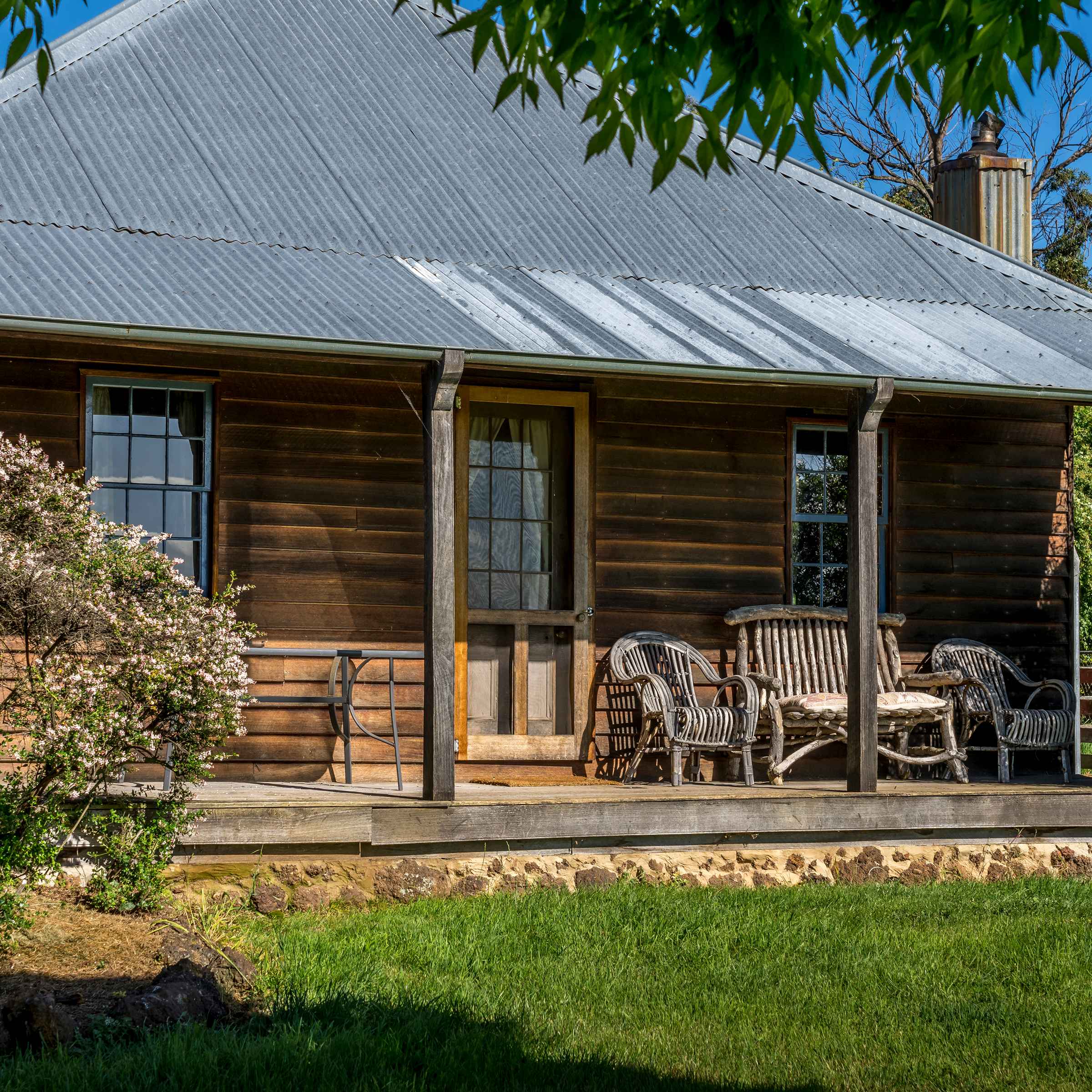 Timber cottage front verandah has several chairs, a timber front door and two windows. Shrubs are at both ends of the verandah and a corrugated iron roof is held by timber verandah posts. A grassed area is in front. Photo: Rob Burnett.