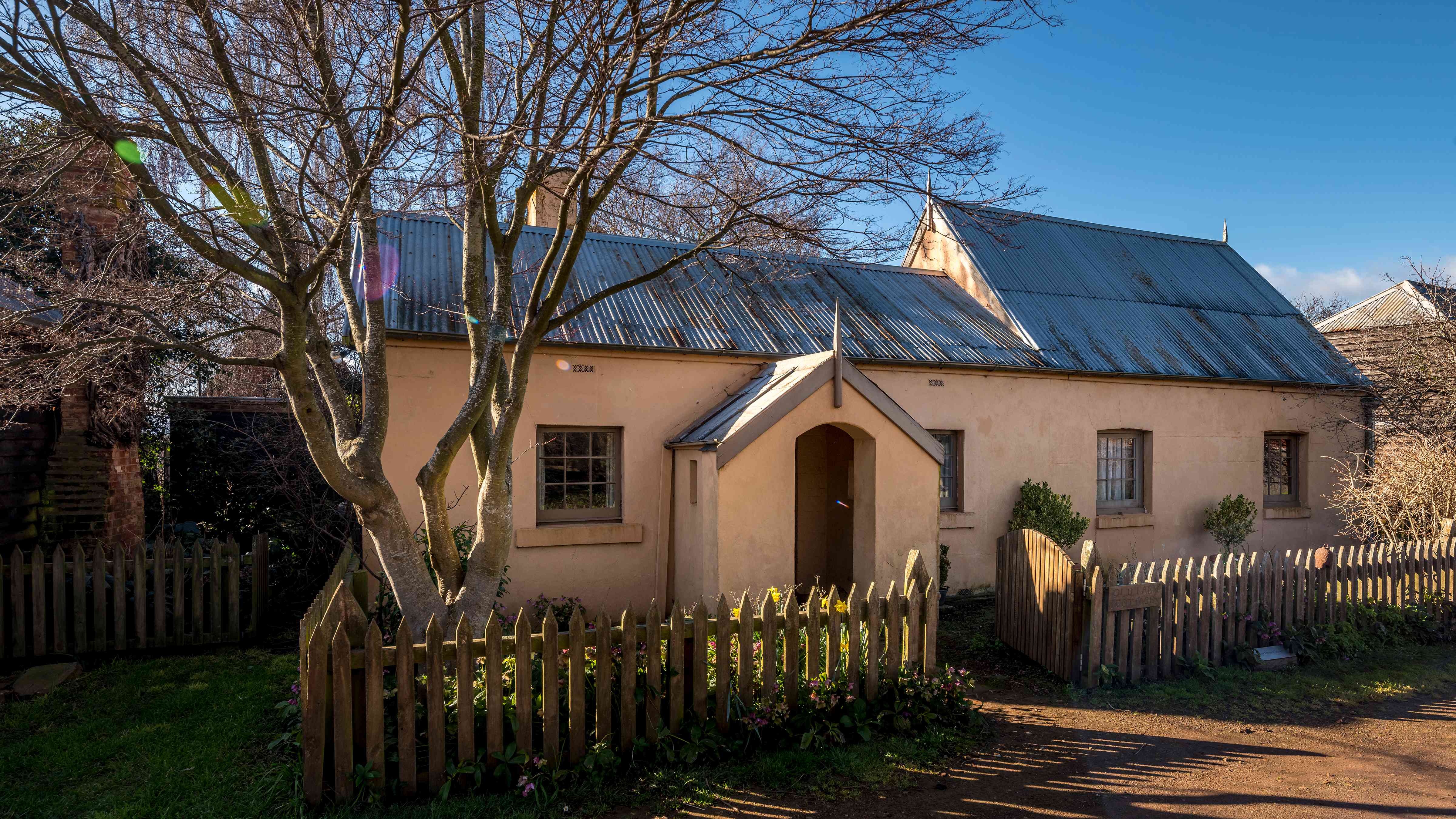 This image was taken in winter as the Japanese elm that stands in the garden does not have any leaves on it. The cottage is pale pink limewashed with a corrugated iron roof. The cottage has two sections, the left is a lower roofed cottage with a window on either side of the front porch and the right section has a steeper pitched roof with two windows. The picket fence runs along the front and the timber gate is open. A bright blue sky is in the background. Photo: Rob Burnett.