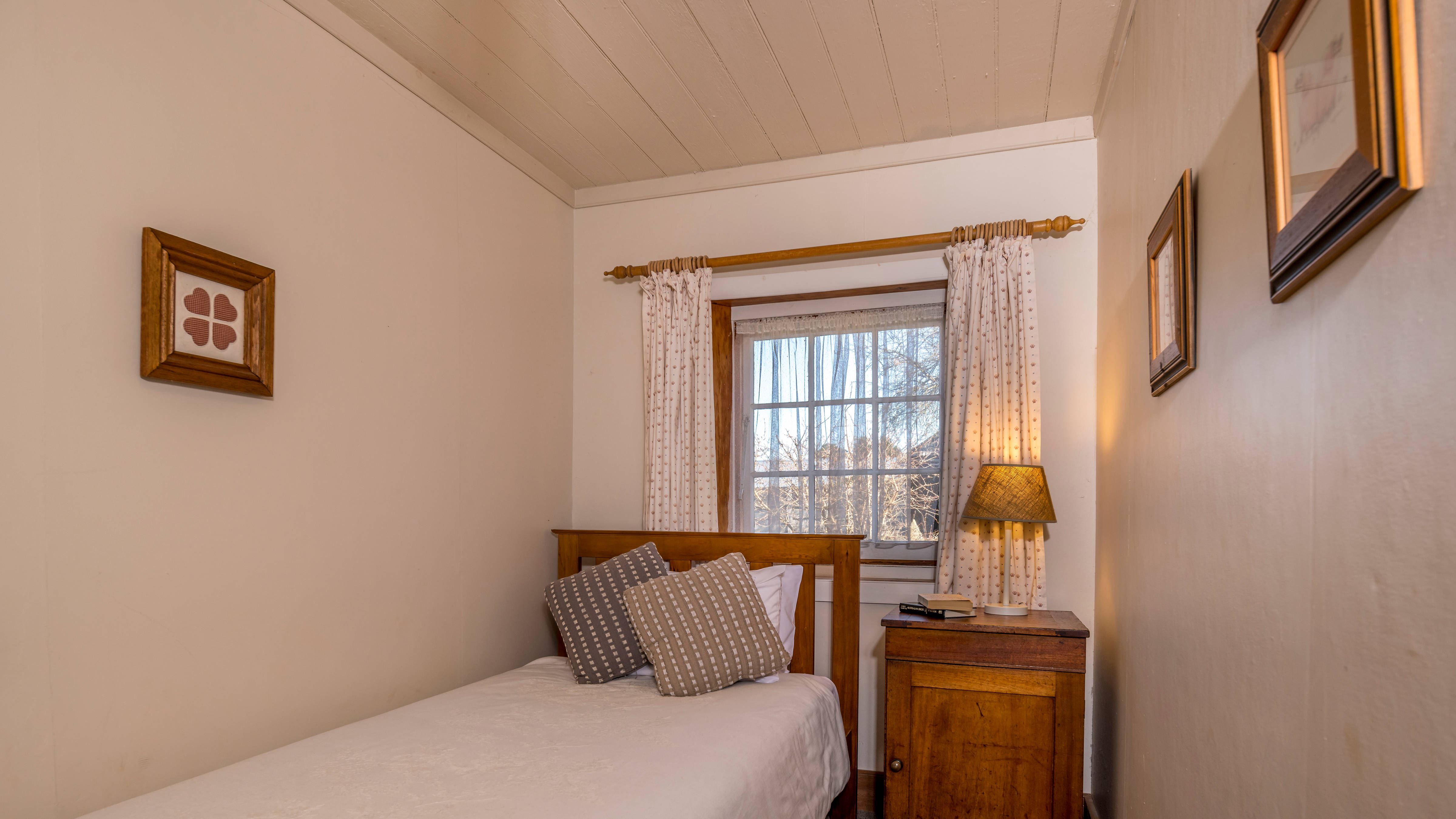 White walls with timber framed pictures hanging lead to the end wall which has a white painted paned window with floral curtains hanging from a timber rod. The timber bed has a fawn quilt cover with two patterned cushions. A cedar bedside cupboard has a bedside lamp and several books on it. Photo: Rob Burnett.