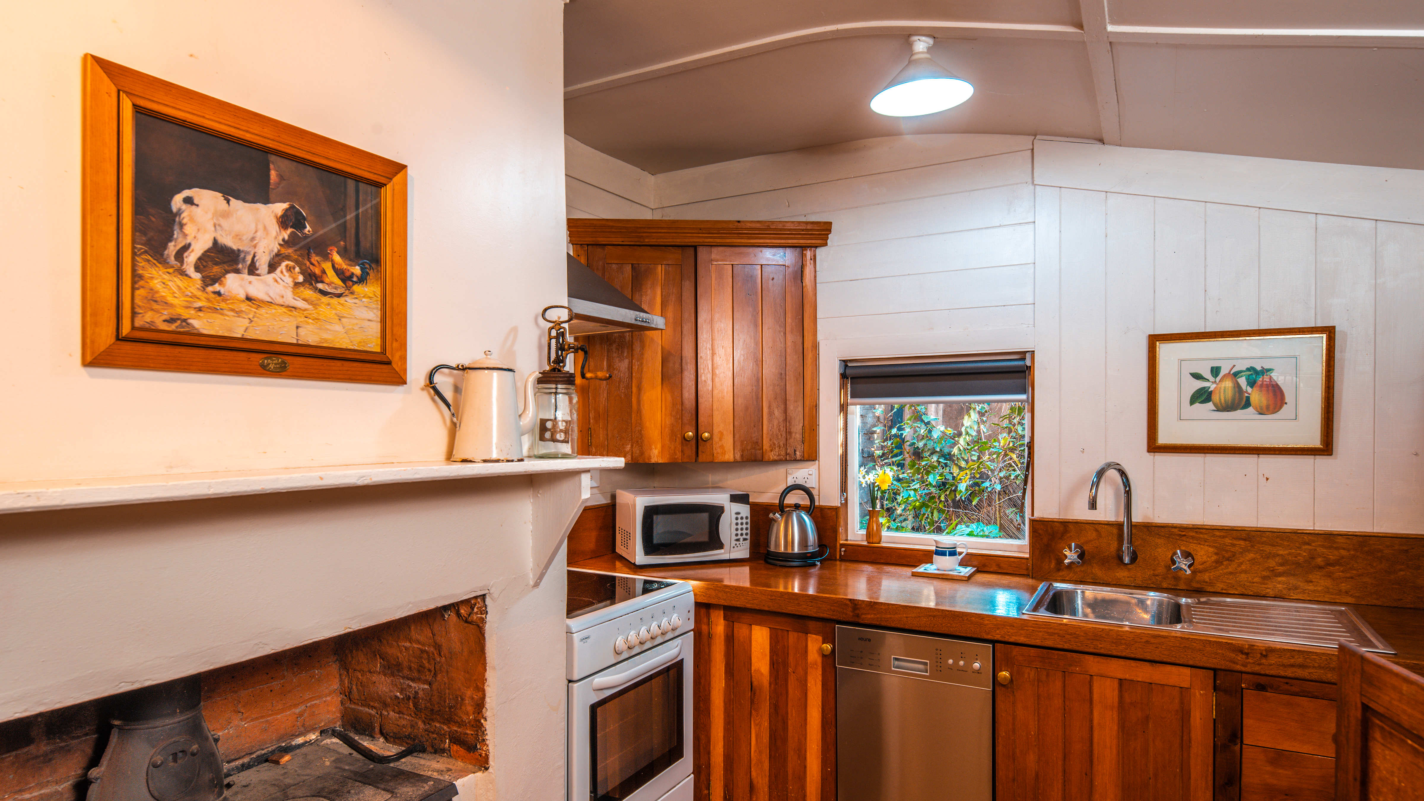 Timber panelled cupboards and a timber bench have a sink with wall taps, microwave, kettle and dishwasher under the bench. A white stove/oven and a white painted timber fire surround stands to the left with a picture of 3 dogs above. A metal jug and old fashioned cream whipper stand on the surround. An old arger stove is in the fireplace. A window is above the bench and looks out to the garden and a picture of pears hangs on the walls. Photo: Rob Burnett.