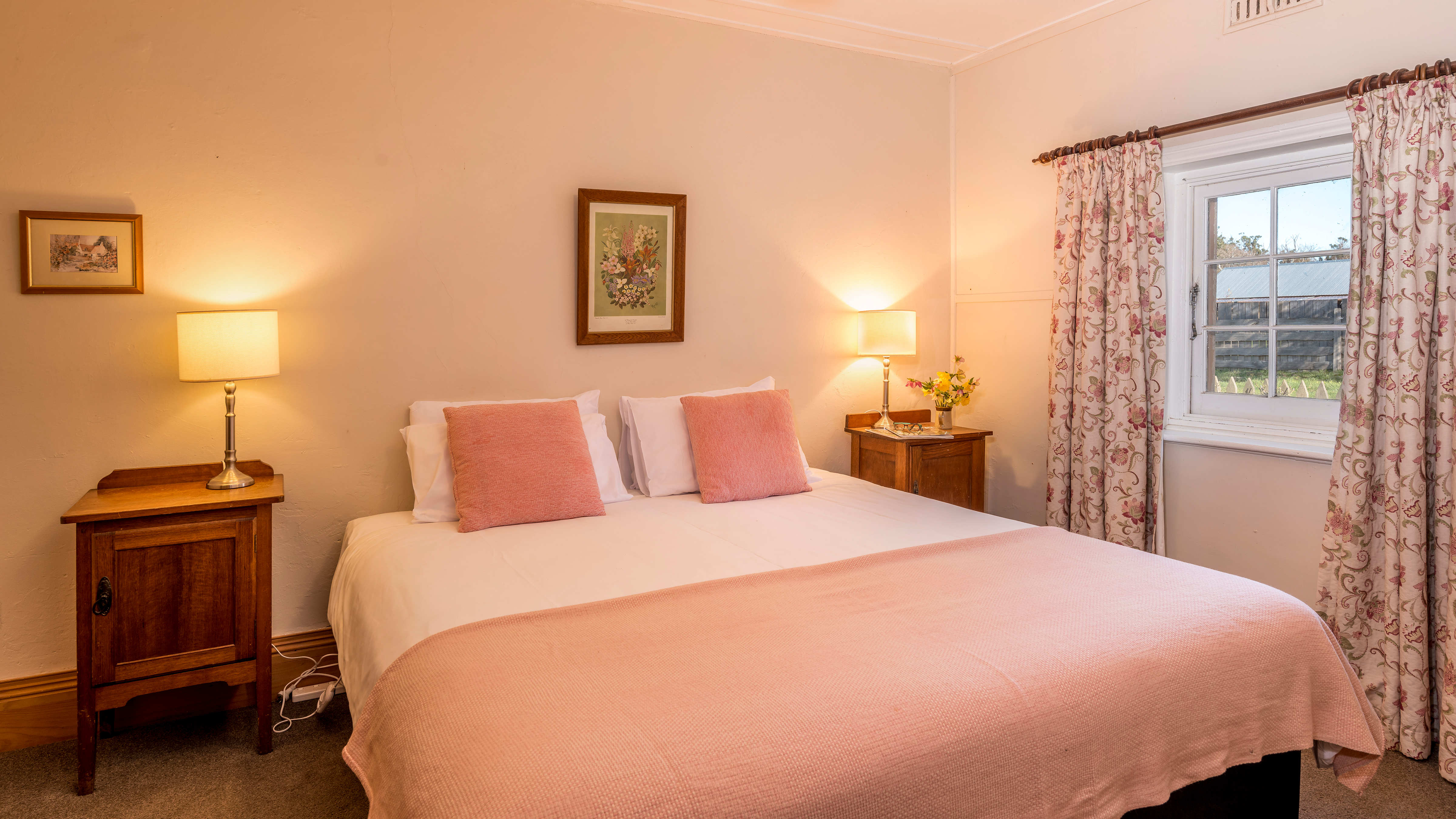 Two bedside cupboards have bedside lamps and a vase of flowers on top. The king bed is made with white linen and a pink rug and pillows. The white painted window has floral curtains hanging on either side and looks out to an old shed. Two timber framed pictures hang on the white walls. Photo: Rob Burnett.