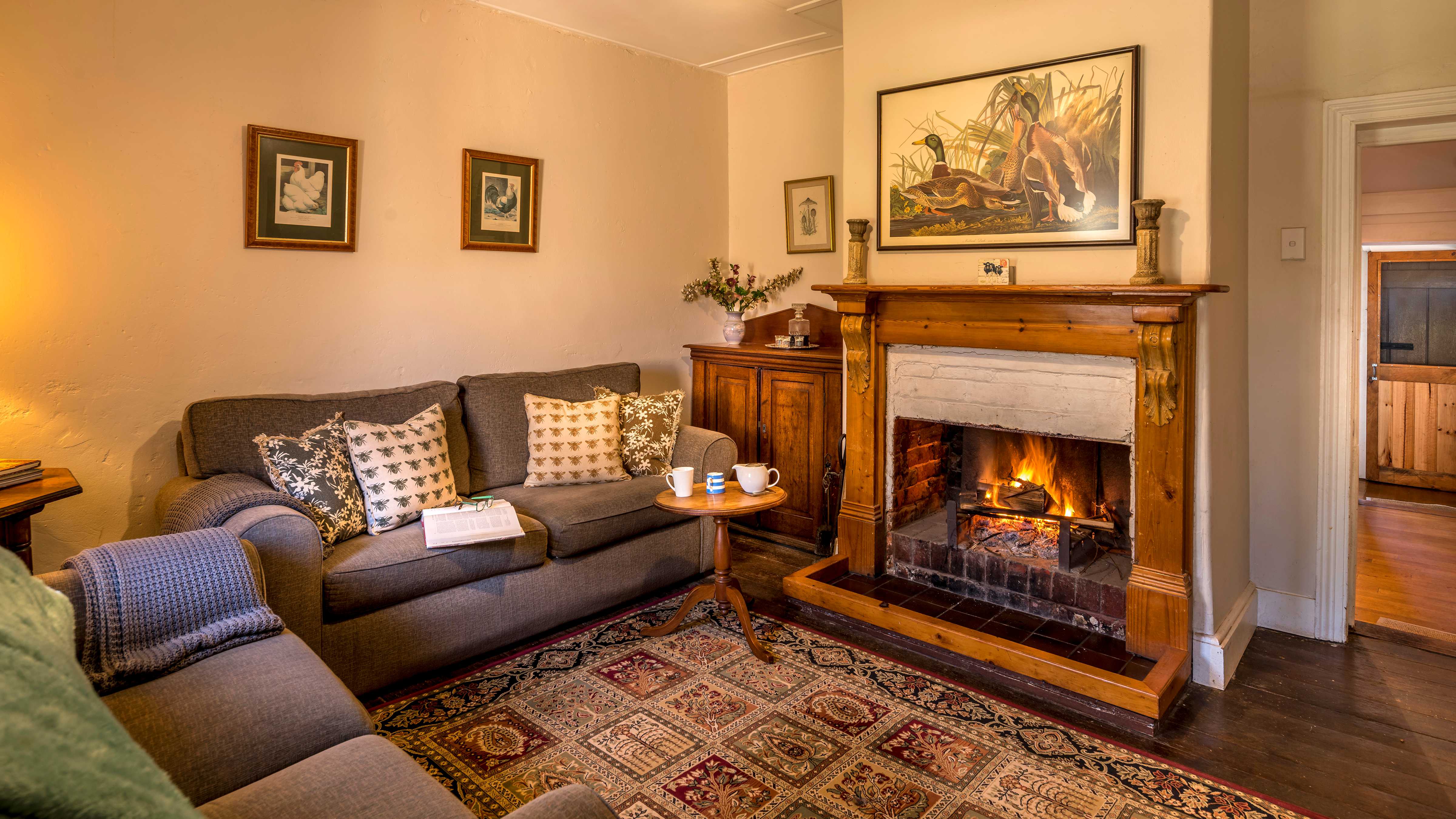 An open log fire is burning in the fireplace which is surrounded by a pine mantlepiece. A large picture which green and brown ducks hangs above the fire and two candle sticks are stand on the mantle. A cedar cupboard has a vase of flowers and a decanter of port with glasses. Two green couches sit at right angles and have patterned green and brown cushions and a grey rug hangs over the arm. A persian style rug in greens and browns covers the timber floor boards. A doorway to the right of the picture leads to another room. Three smaller pictures hang on the white painted walls. Photo: Rob Burnett.