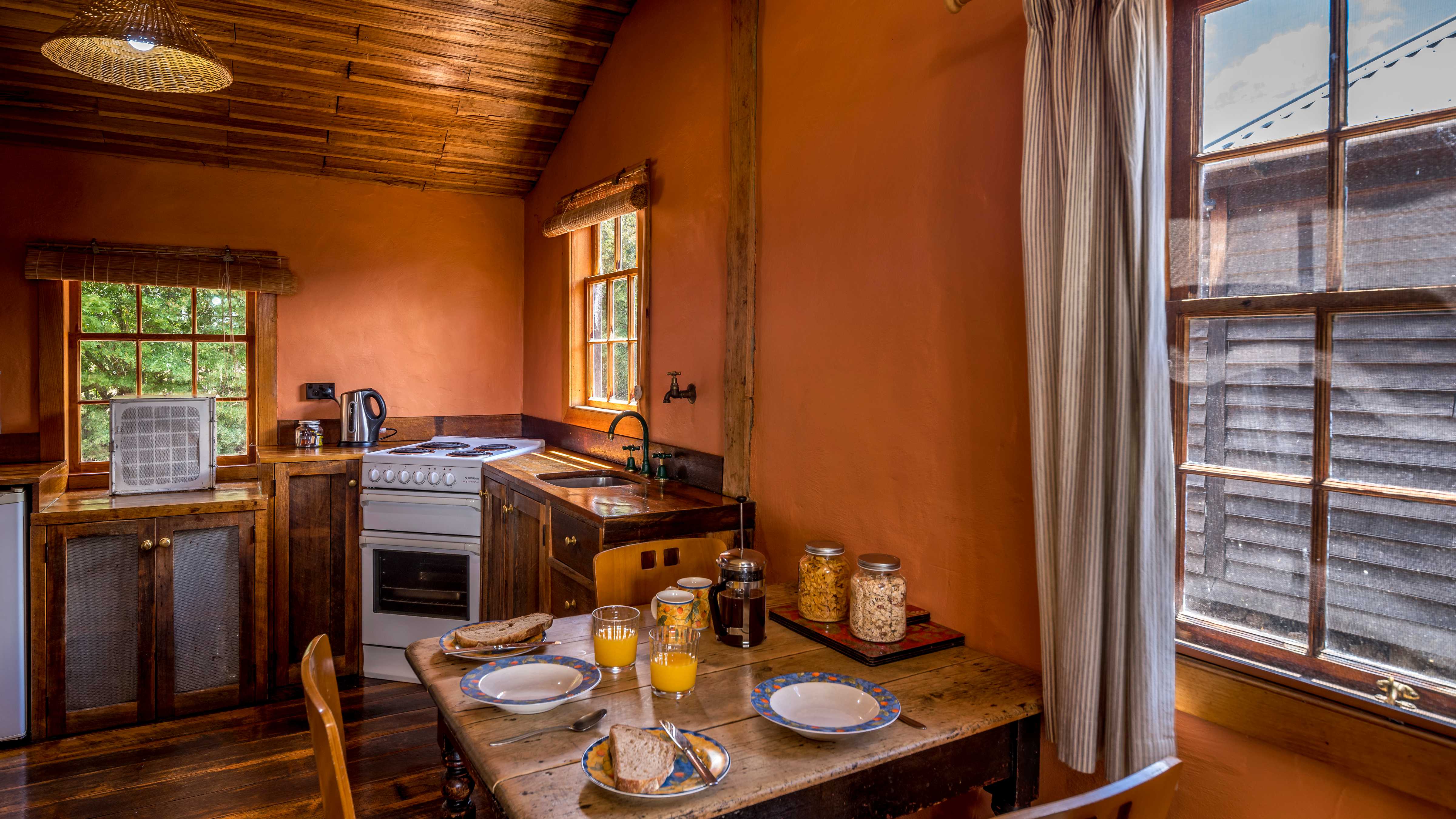 The open plan farmhouse kitchen has timber cupboards and table and chairs are bordered by georgian paned windows and terracotta painted walls. The table is laid with breakfast provisions including jars of cereal, glasses of orange juice, bowls and plates with toast and a coffee plunger. Photo: Rob Burnett.