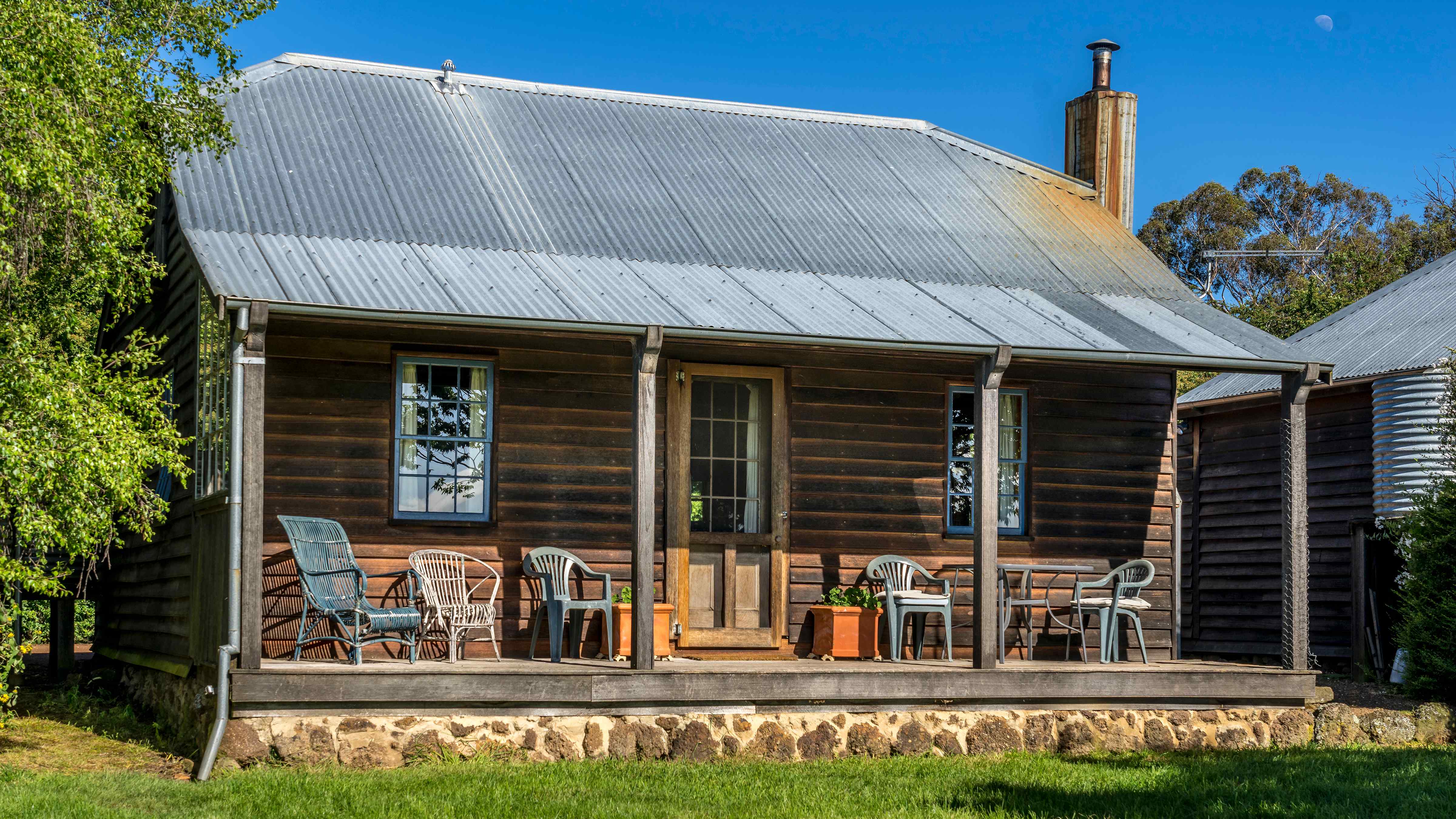 The verandah has multiple chairs and two terracotta pots on either side of the front door. The weatherboard front wall has two windows and a front door in the middle. A corrugated roof has a chiney to the right hand side. Rock foundations support the verandah and grass is in the foreground. Photo: Rob Burnett.