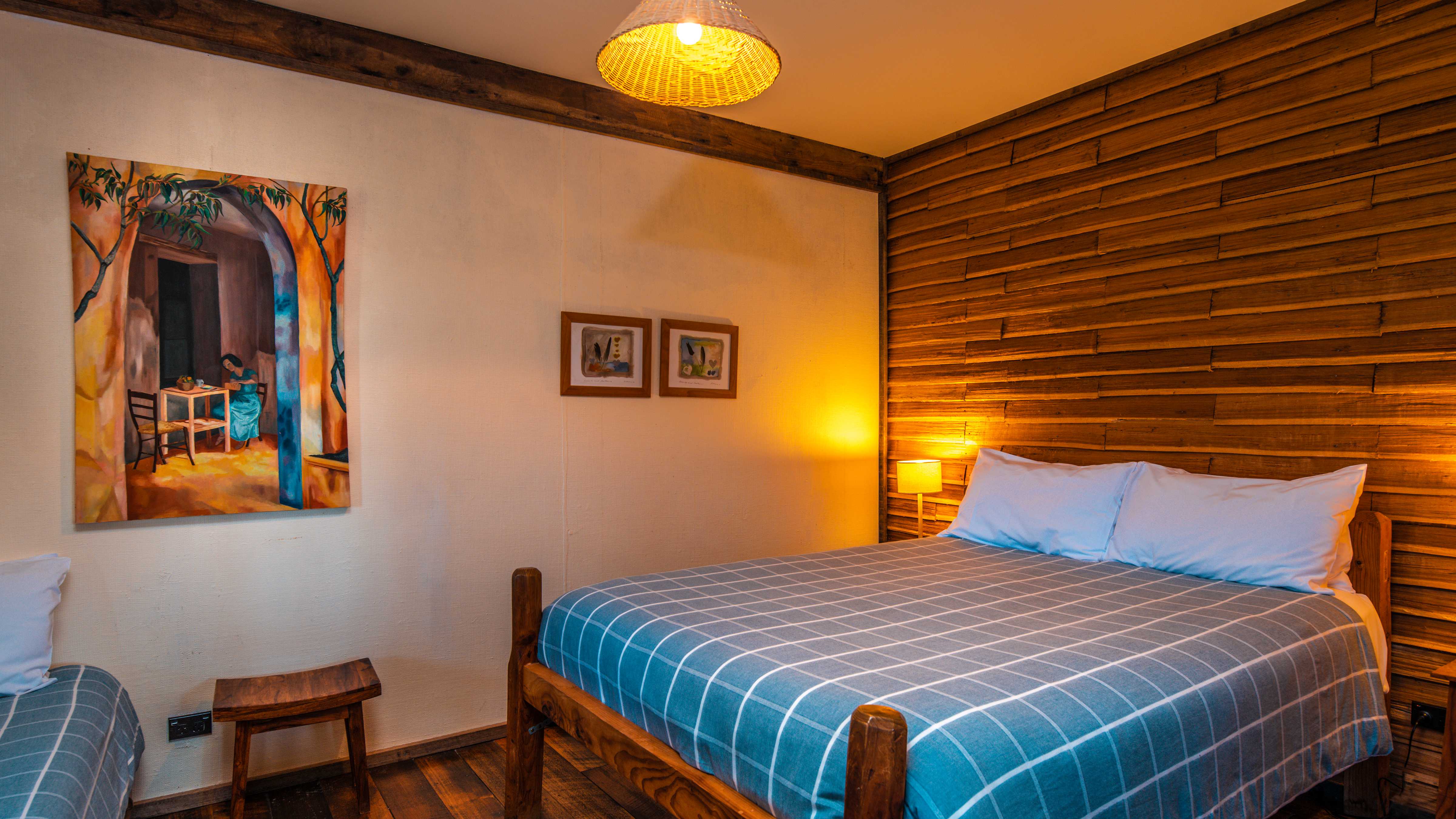 A timber queen bed sits to the right of the room with a blue and white checked quilt and white pillows. The wall behind the bed is horizontal timber palings and the other wall is white painted hessian. A large painting of a woman sitting at a table in blues and yellows hangs on the white wall and two smaller prints are alongside. A lamp that is turned on sits on the far side of the bed and the main light is also on. A small timber table sits beside another single bed on the left. Photo: Rob Burnett.