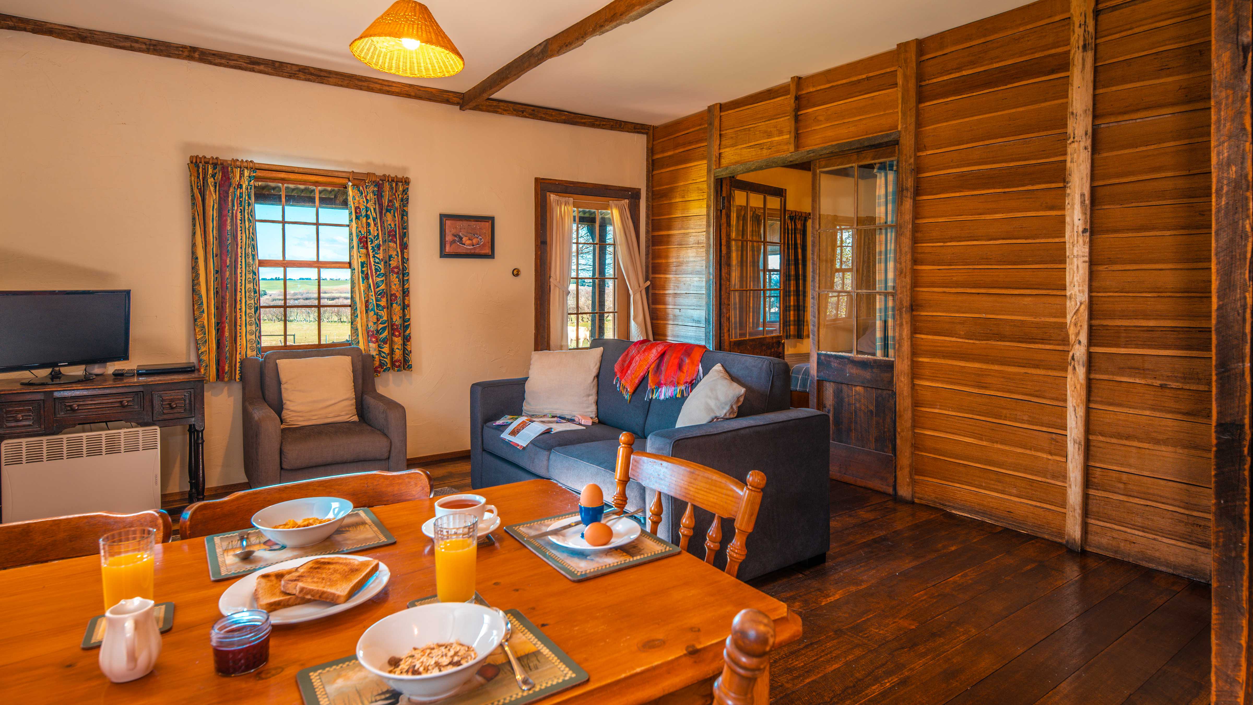 A timber table is set with breakfast which includes bowls of cereal, glasses of orange juice, toast on a plate, boiled eggs in an egg cup and a cup of tea. A dark coloured lounge has light colour cushions and a throw on it and a single lounge chair sits to the left where there is also a TV on a timber side table and heater. A window has pattern curtains hanging on either side and looks out to farmland. There are white painted walls and a horizontal timber paling wall with a timber and glass door open to the bedroom. Timber floor boards are to the right of the image. Photo: Rob Burnett.