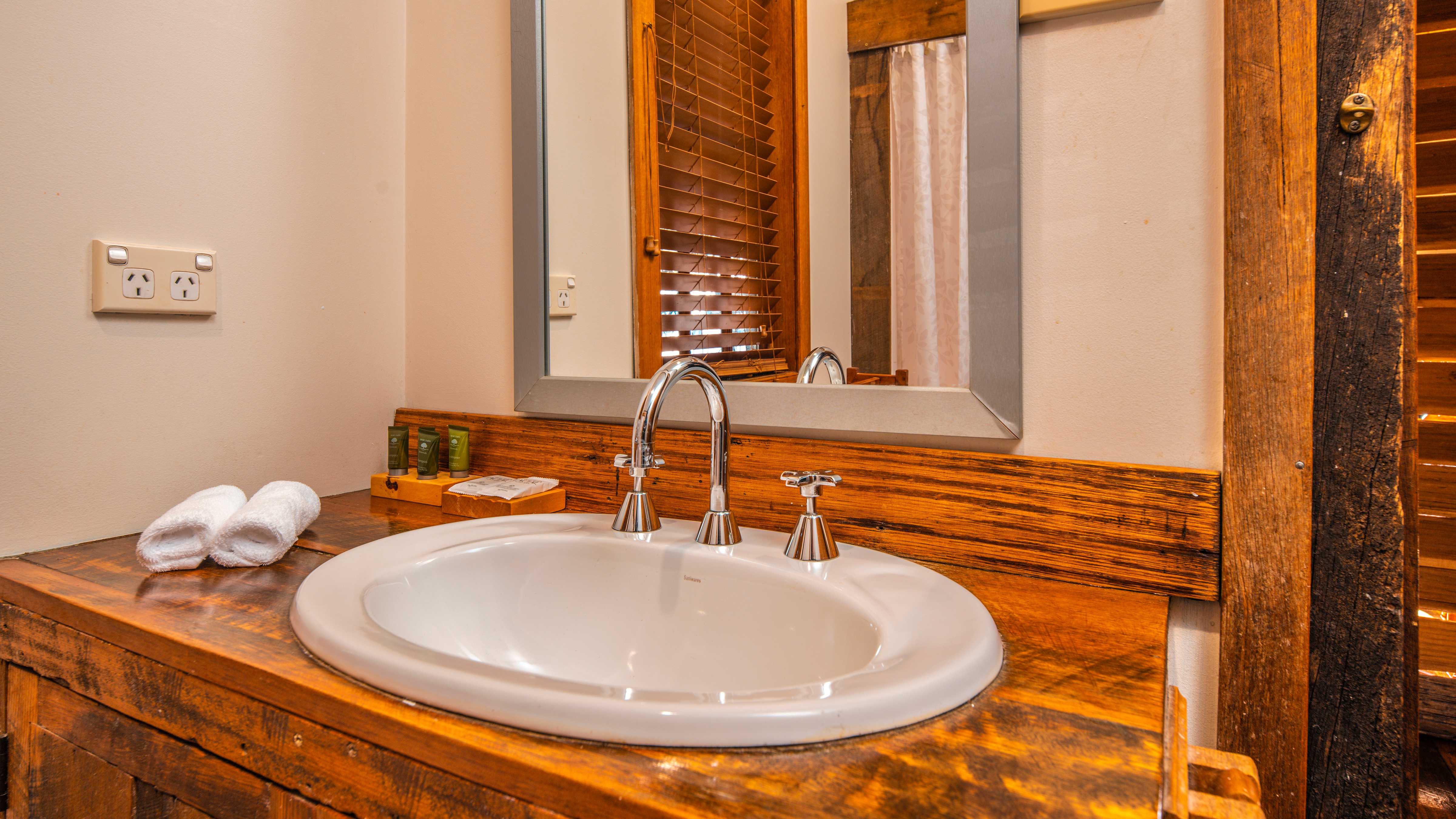 A rustic timber vanity has a white oval basin with chrome taps and spout. Two white face cloths are rolled to the left with a power point on the wall. A chrome framed mirror hangs on the white painted wall behind the basin. Photo: Rob Burnett.