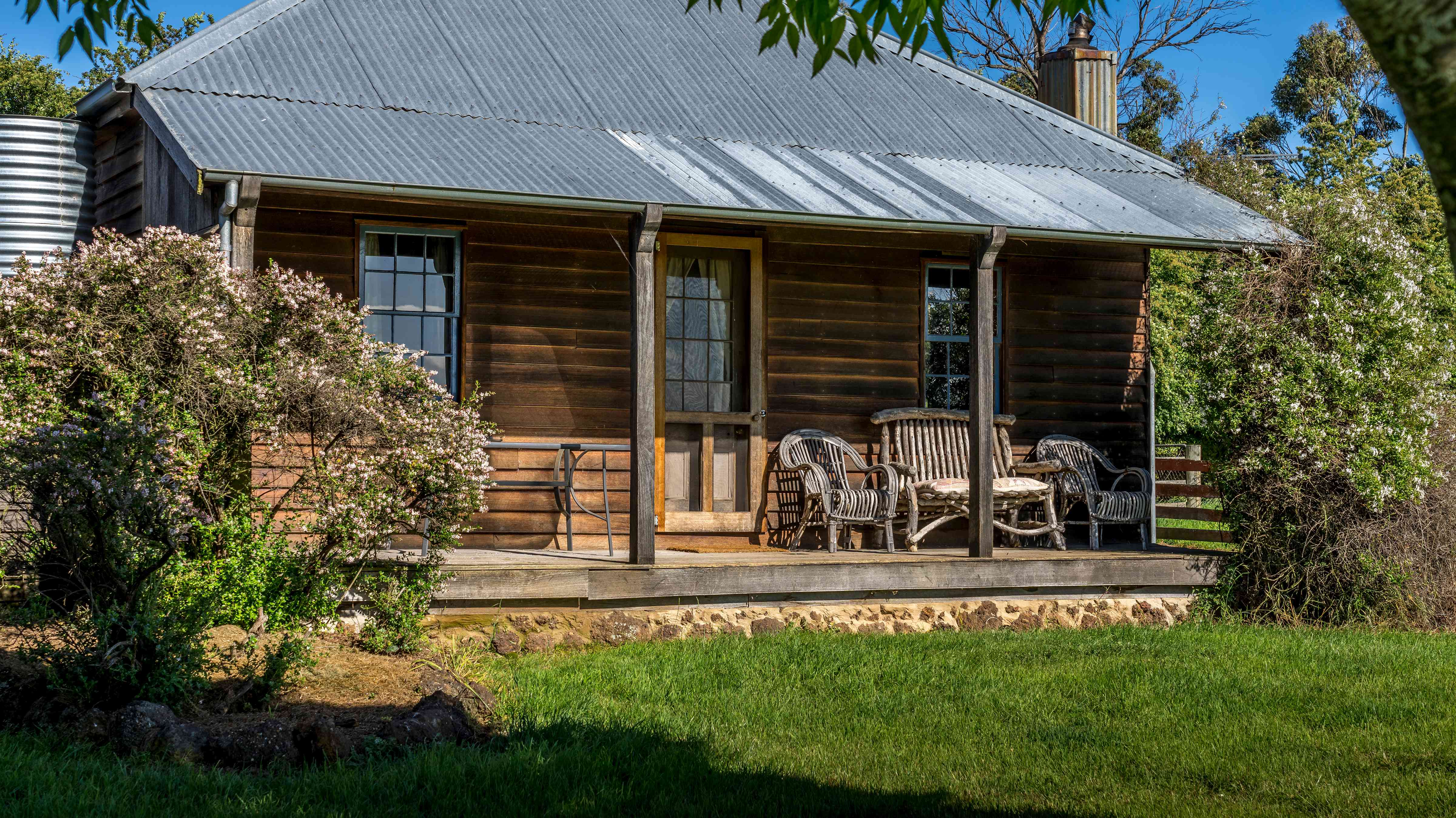 Timber cottage front verandah has several chairs, a timber front door and two windows. Shrubs are at both ends of the verandah and a corrugated iron roof is held by timber verandah posts. A grassed area is in front. Photo: Rob Burnett.