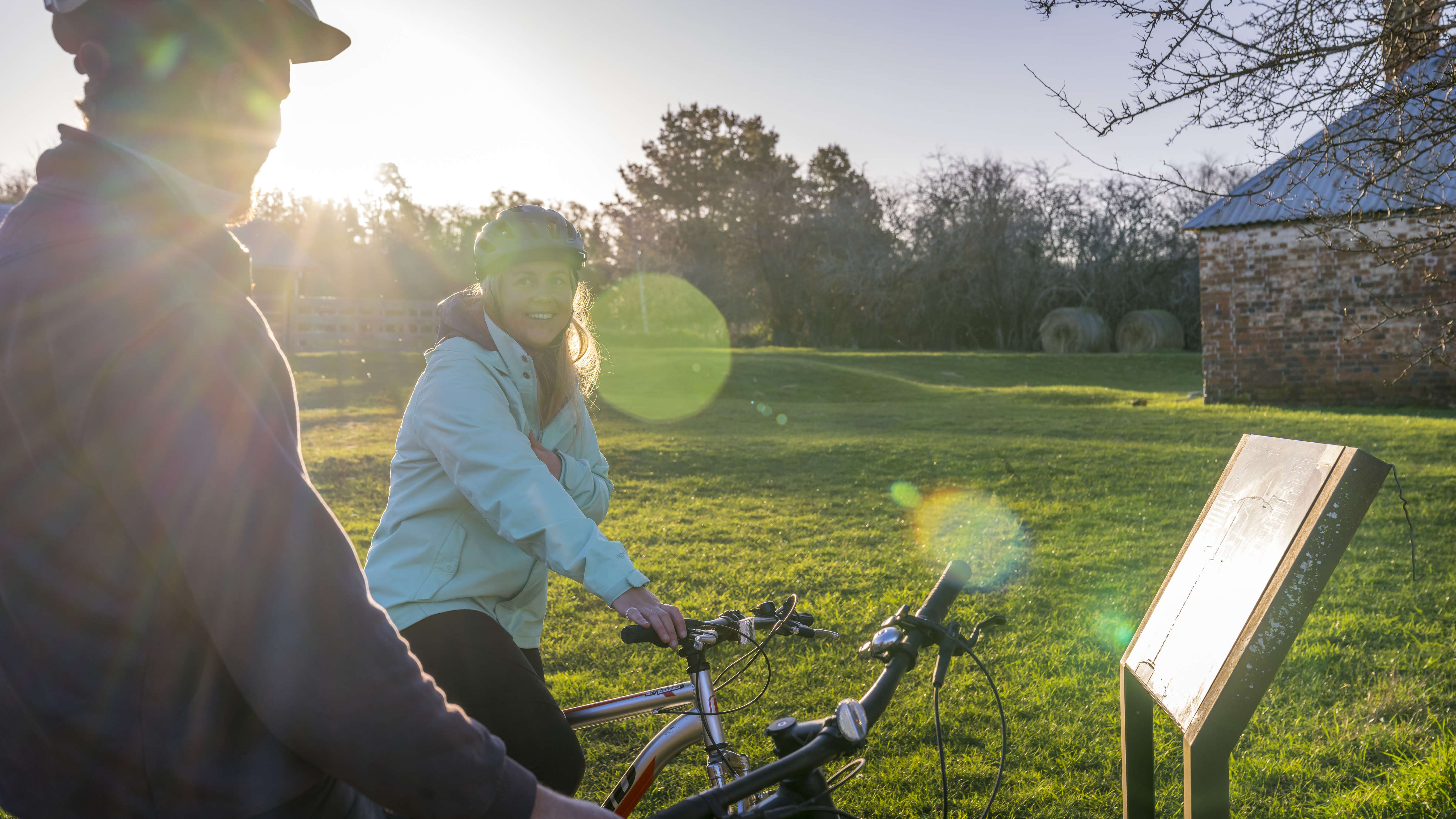 Two cyclists read an information sign at the start of the Convict Trail. The corner of the blacksmiths shop is in the background surrounded by green grass and the sun behind the hedgerows. Photo: Rob Burnett.