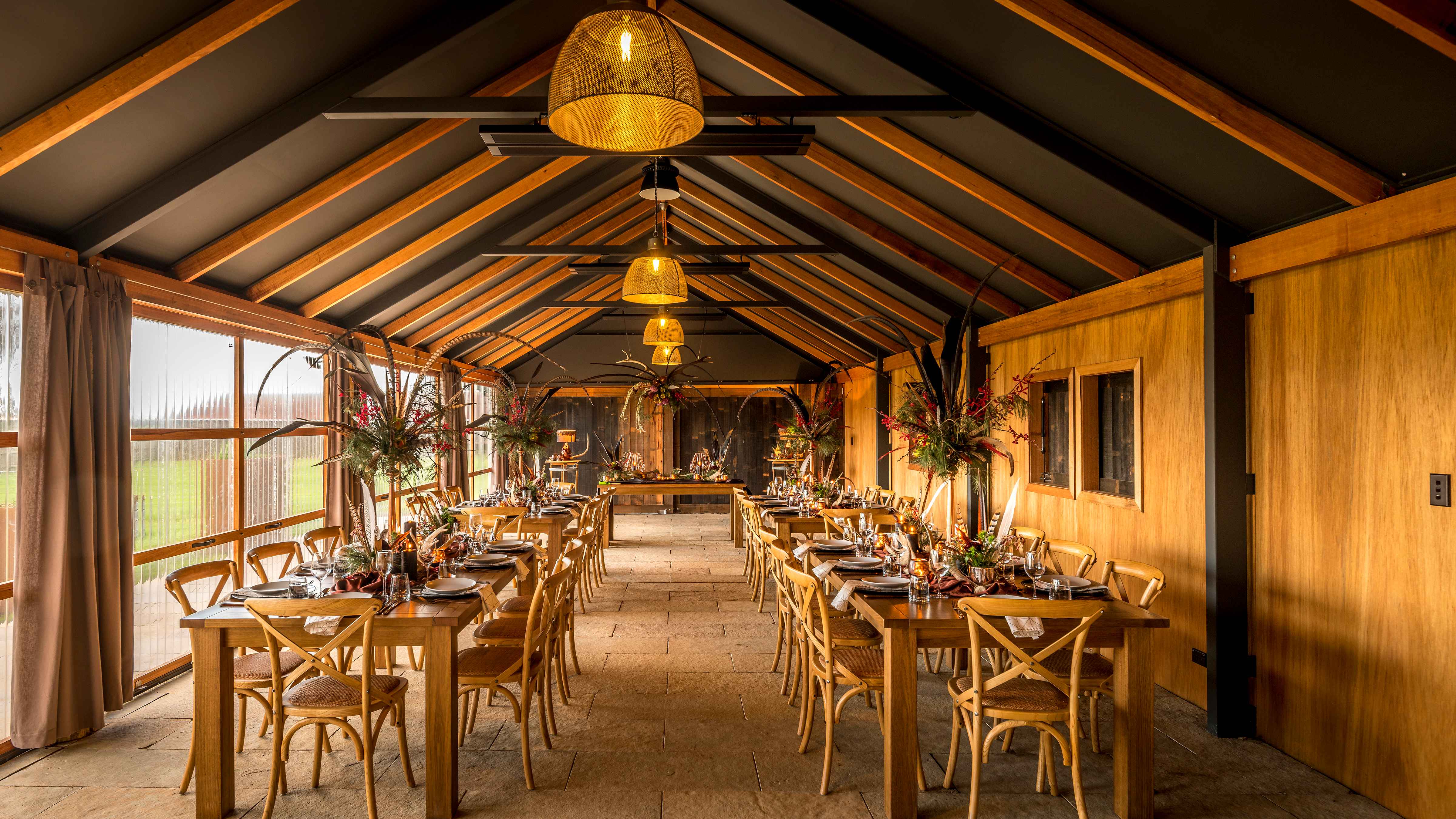 Long wooden tables are set in The Archery with crockery, cutlery and large floral arrangements. Wooden cross back chairs are placed at the tables and four pendant lights hang from the ceiling. The left wall is made up of clear doors that are closed. The floor is limestone pavers and exposed rafters line the ceiling. Photo: Rob Burnett.