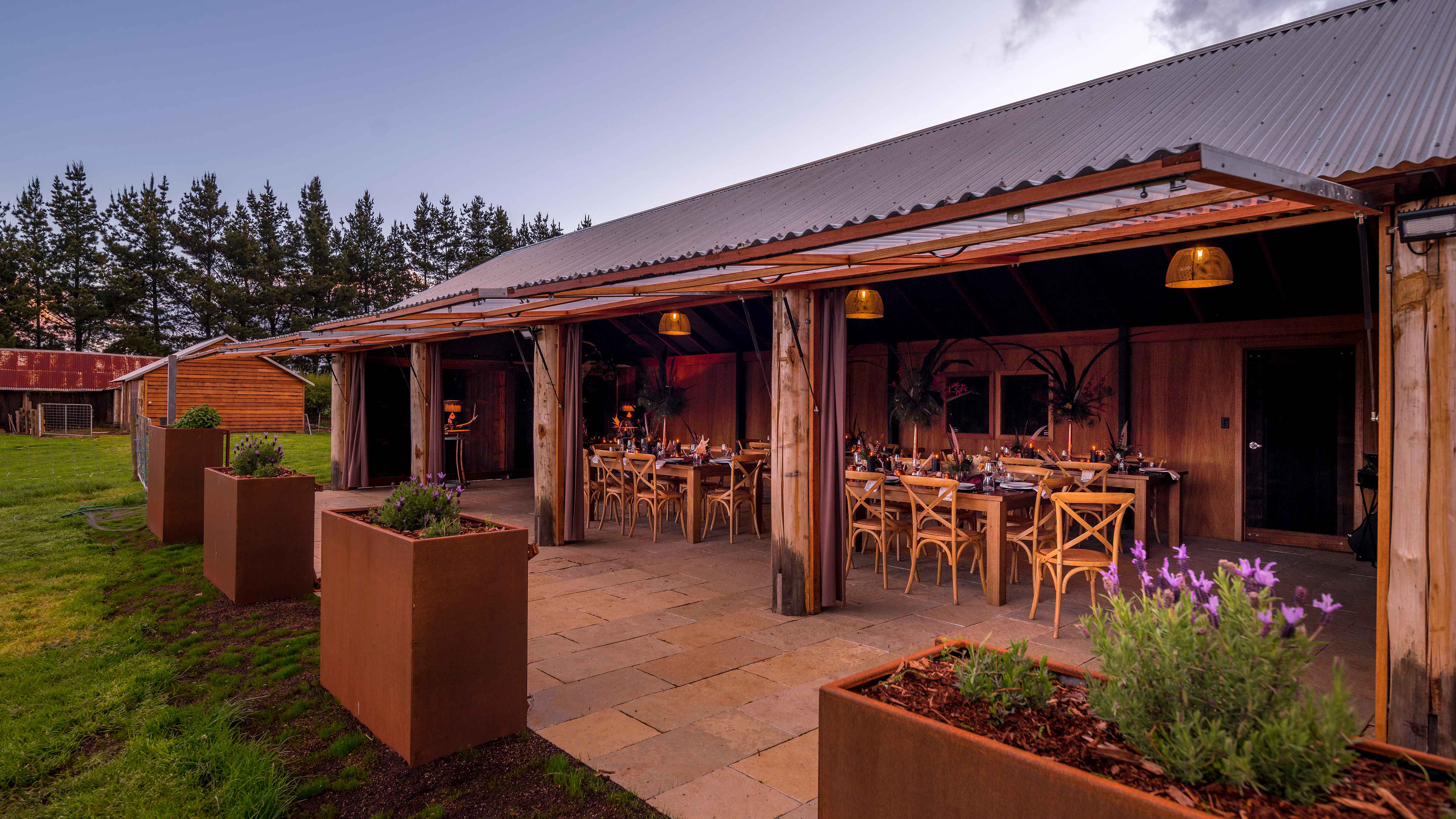 The vertical corrugated laserlight doors are open on the Archery building. Inside are tables set with timber cross back chairs and decorations. Iron planter boxes border the limestone pavers that run along the front and continue inside. There is a row of pine trees in the distance behind another timber building. Photo: Rob Burnett.