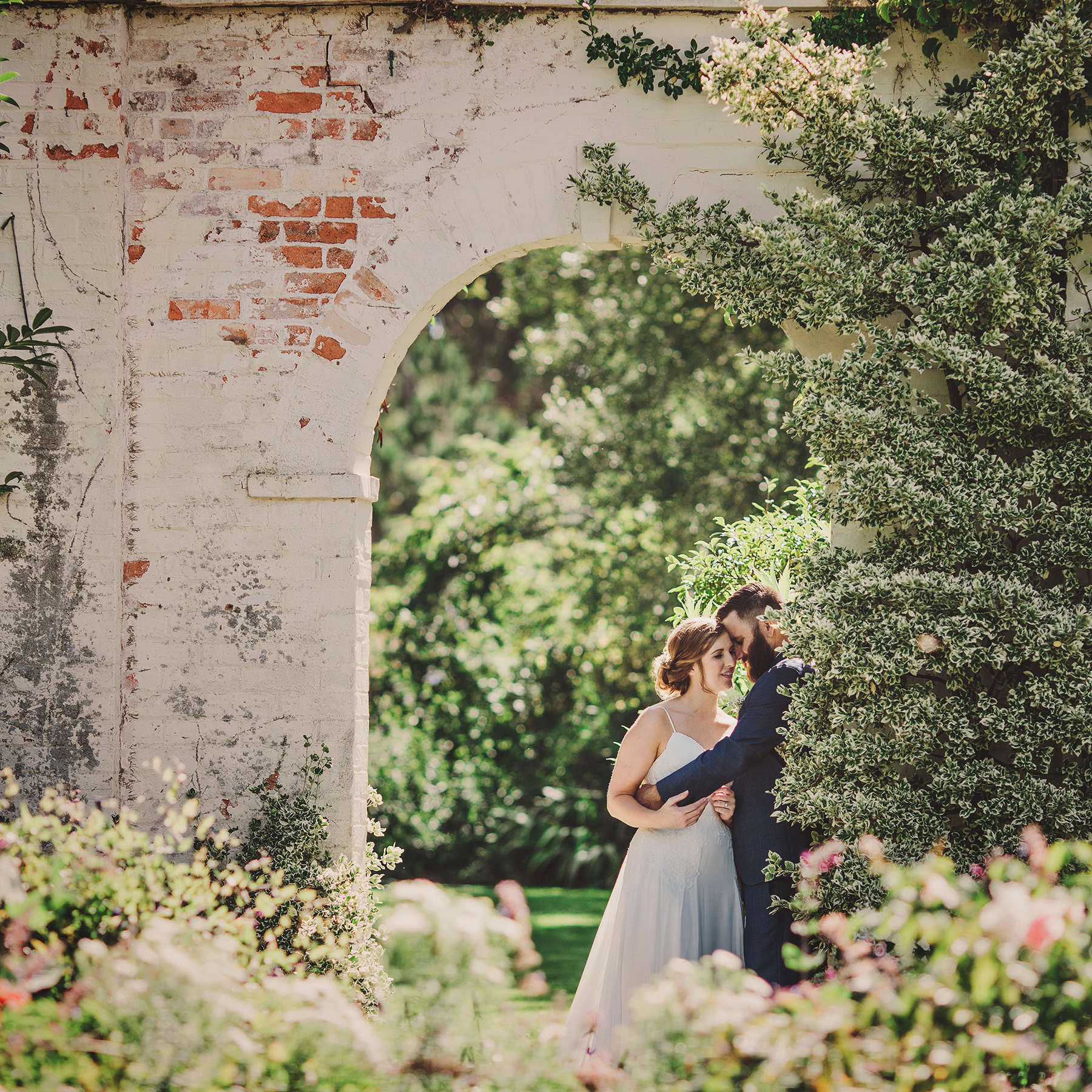 A bride and groom embrace under a white brick archway surrounded by climbing plants and flowers. Photo: Astrid Simone.