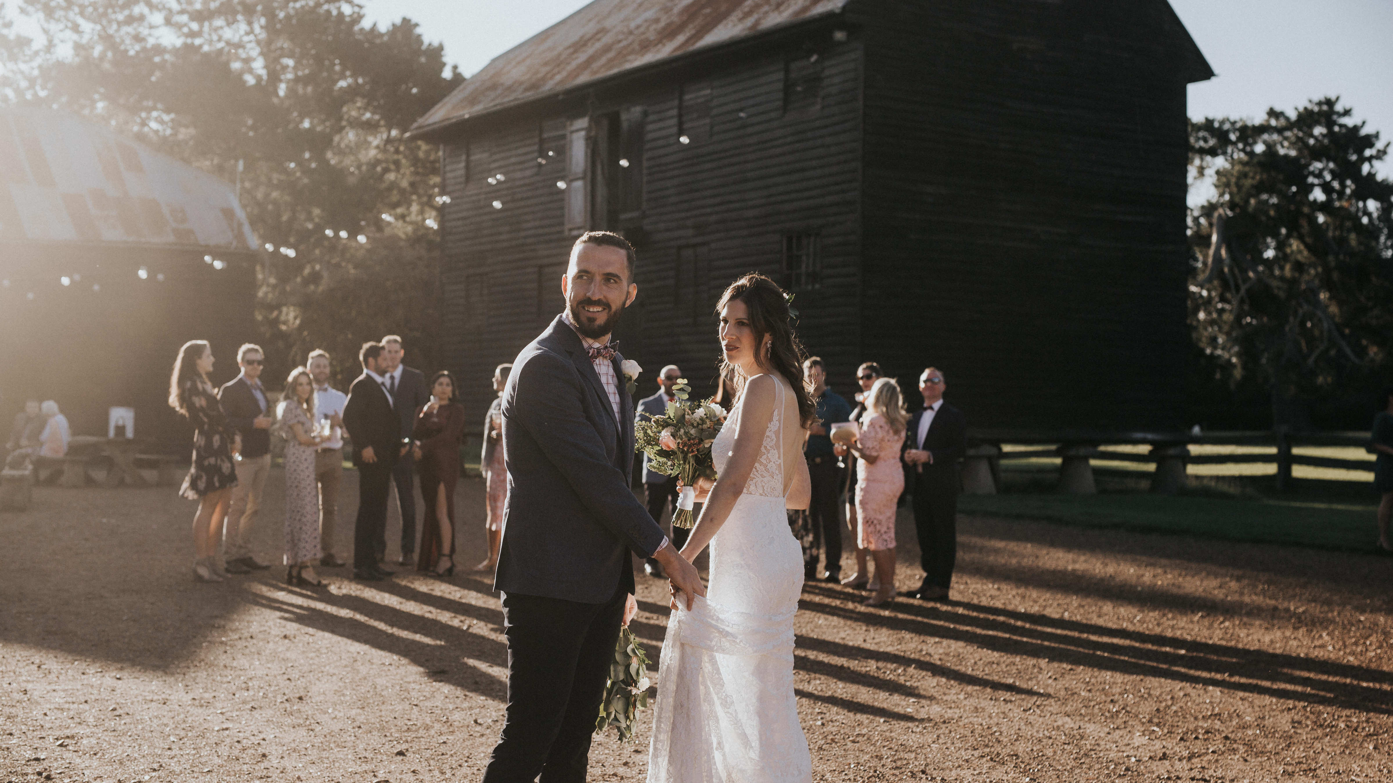 A bride and groom turn to look past the camera as guests mingle behind them in the quadrangle. The timber Pillar Granary stands behind the guests with festoon lights draped from it. Photo: Cassie Sullivan.