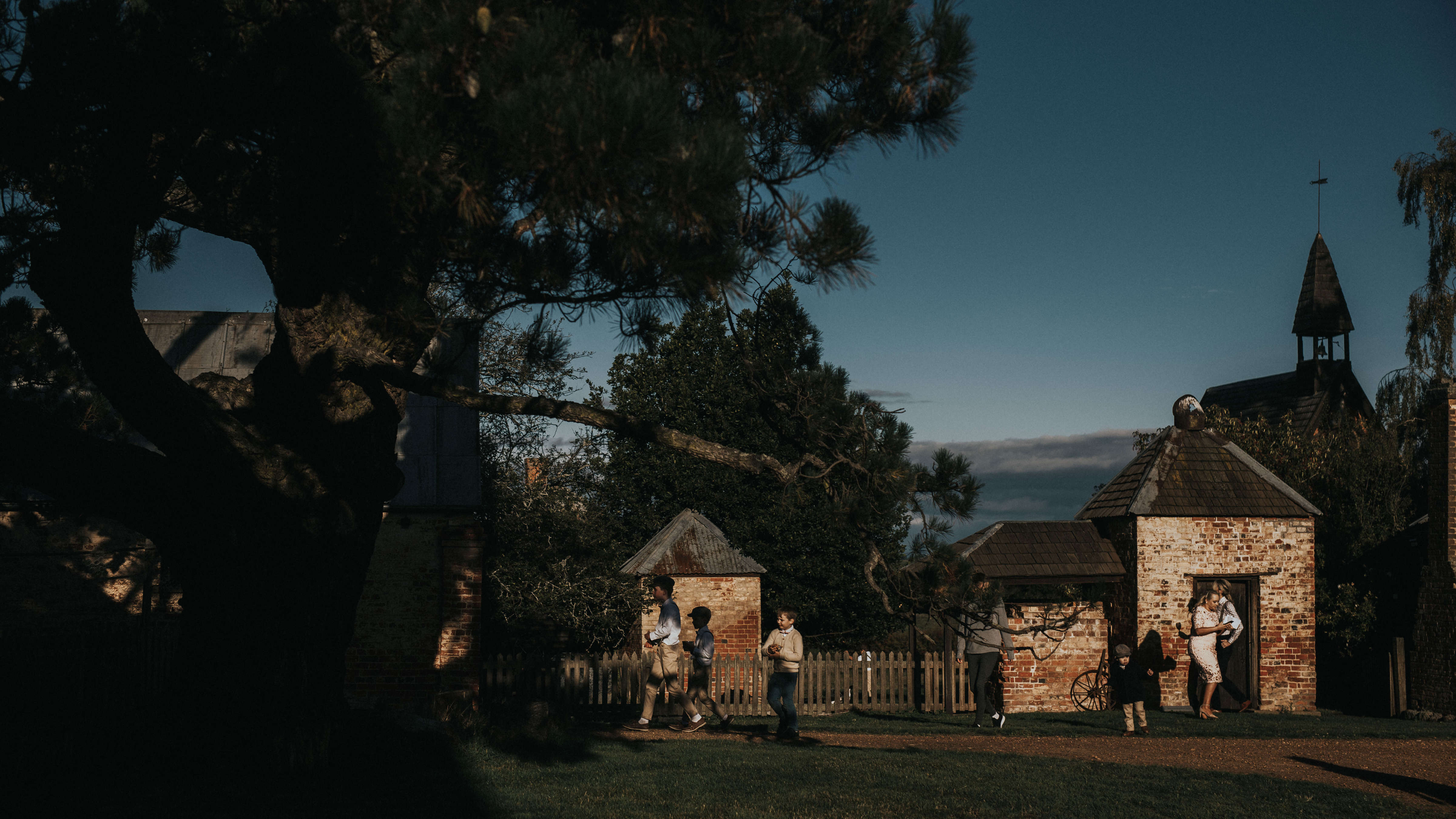 Children walk in front of the smokehouse and picket fence into the shadows of the pine tree. The Chapel can be seen rising up behind the brick smokehouse. Photo: Cassie Sullivan.