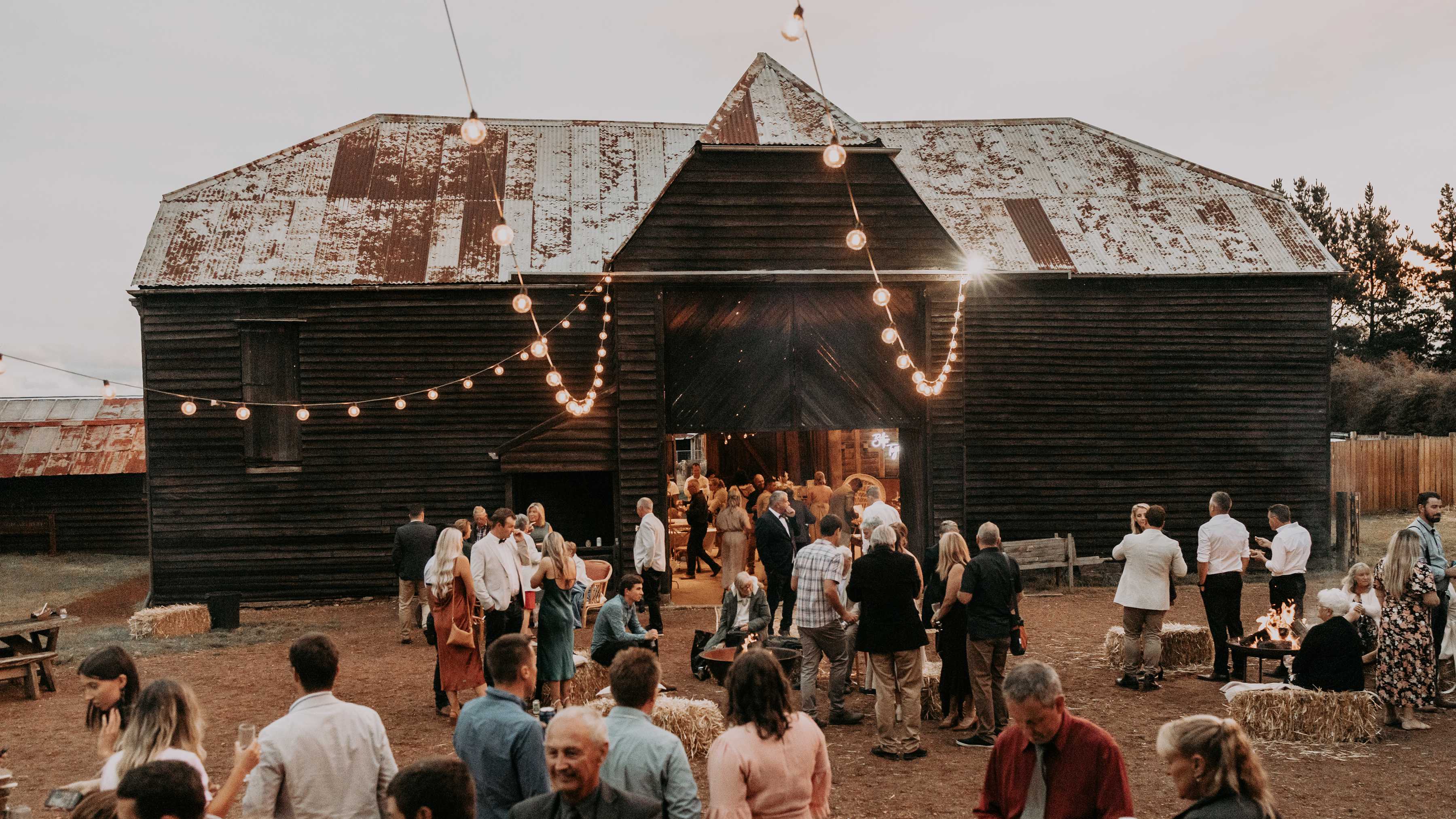 The Sussex timber barn and quadrangle is set for an outdoor wedding reception. Hay bales, fire pits and festoon lights decorate the area along with timber outdoor tables. Guests are mingling outside and inside the Barn. Photo: Tiarne Shaw Photography.